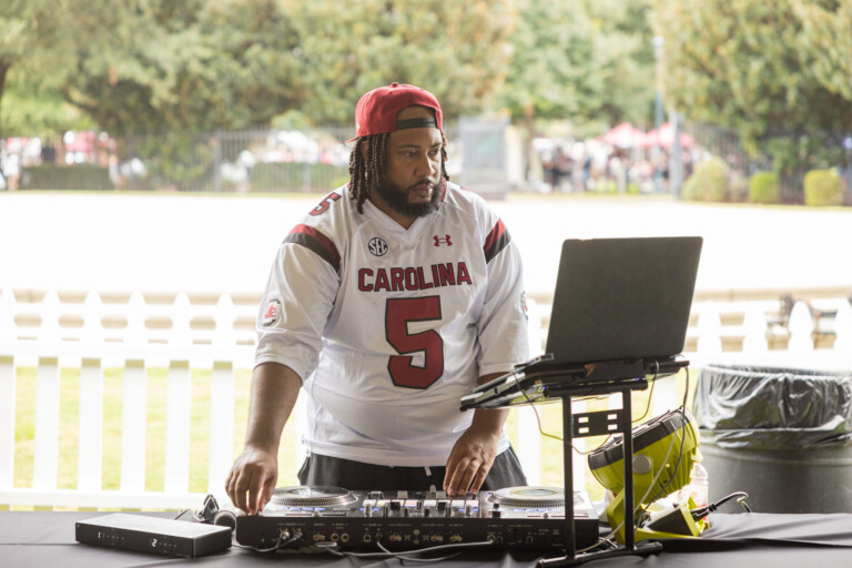 A DJ in a white South Carolina jersey with the number 5 mixes music at a table outdoors, with a laptop and turntables.