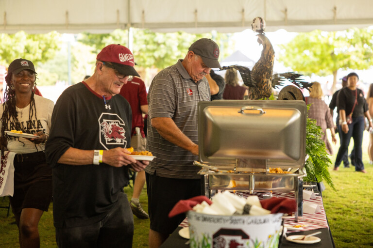 Two men serving themselves food from a buffet under a tent, with a decorative Gamecock statue in the background.