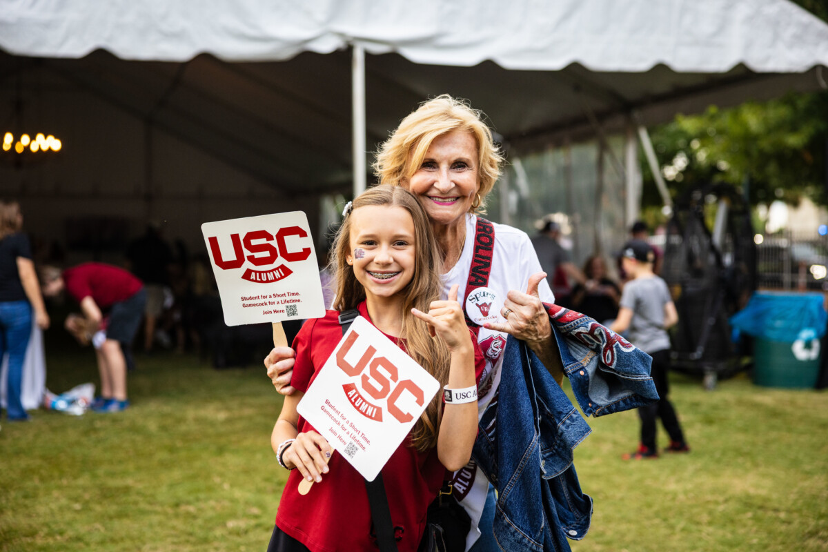 A young girl and an older woman pose together, both smiling and holding USC Alumni signs at a festive outdoor gathering.
