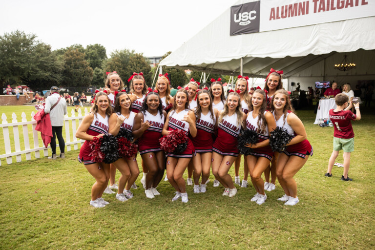 A group of cheerleaders in red and white uniforms poses together outdoors, smiling at the camera, with a tailgating event in the background.
