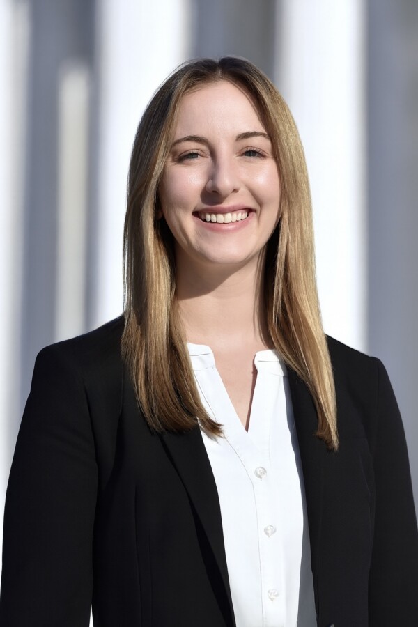 A smiling young woman with long, straight hair wearing a black blazer and a white blouse, standing outdoors against a light background.