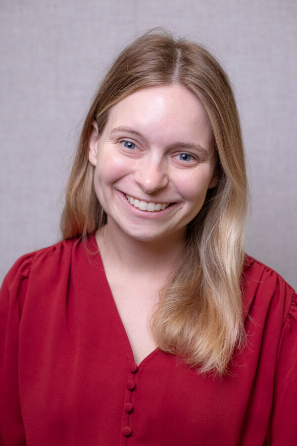 A smiling woman with long blonde hair wearing a red blouse, against a neutral gray background.