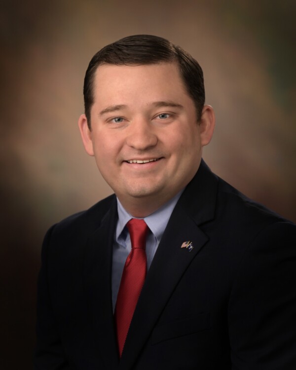A smiling man in a dark suit and red tie, posed against a blurred pastel background.