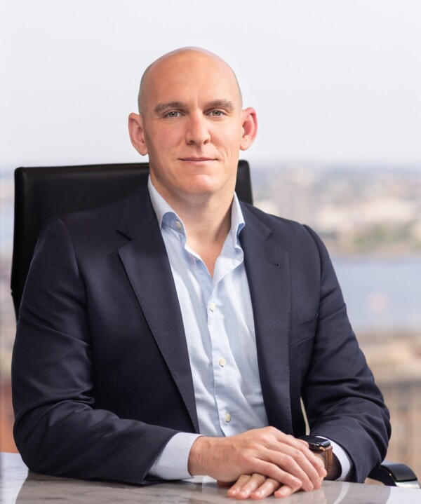 A bald man in a dark suit sits at a desk, looking confidently at the camera, with a city skyline visible in the background.