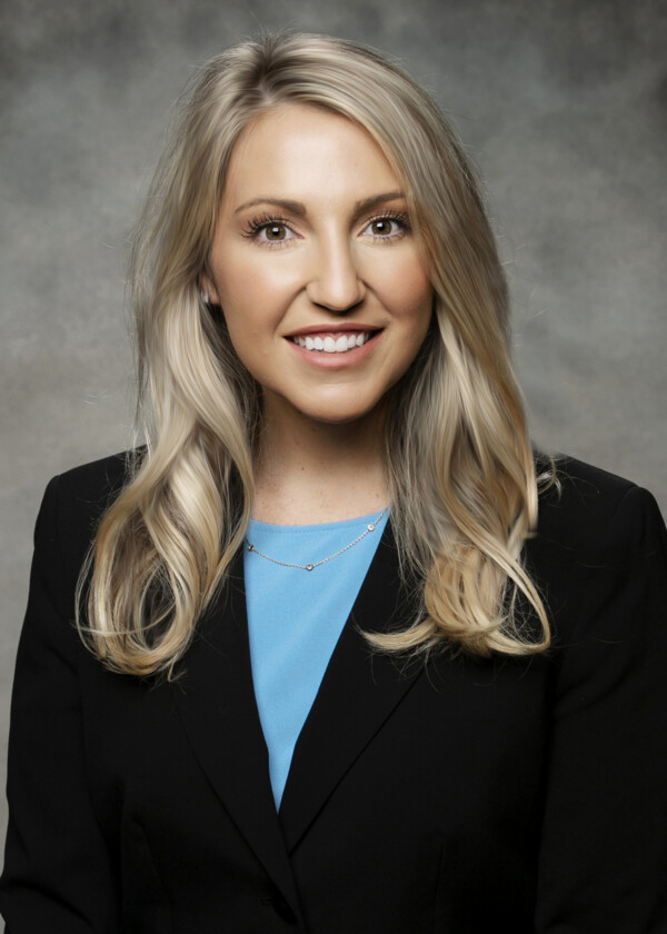 Smiling woman with long, blonde hair wearing a black blazer and blue top against a gray background.