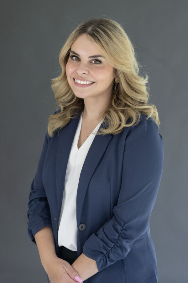 A young woman with wavy blonde hair smiles confidently while wearing a navy blazer and a white blouse, posed against a neutral background.