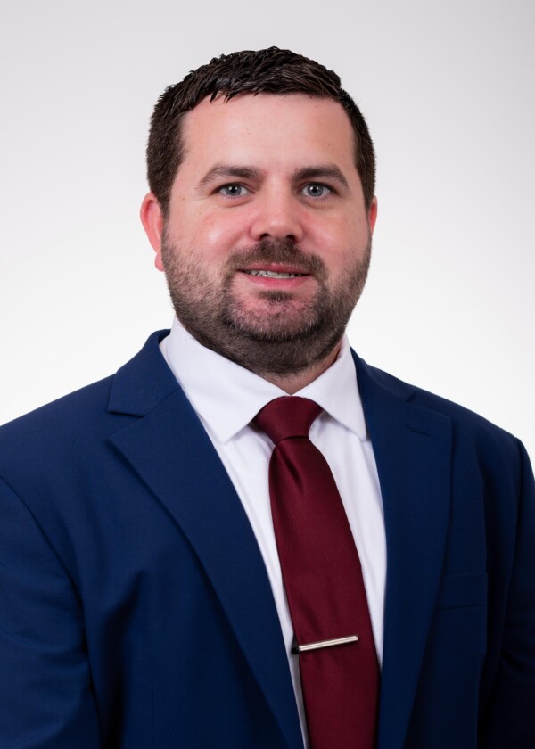 A man in a blue suit and red tie poses for the camera, smiling with a neutral background.