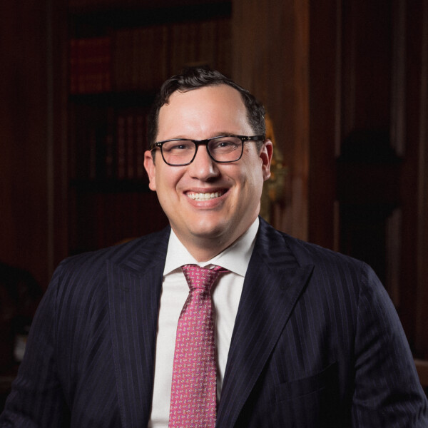 A man in a pinstripe suit sits confidently on a wooden desk with books, smiling in a richly decorated room.