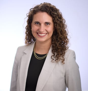 A smiling woman with curly hair wearing a light gray blazer and layered gold necklaces against a soft, light background.