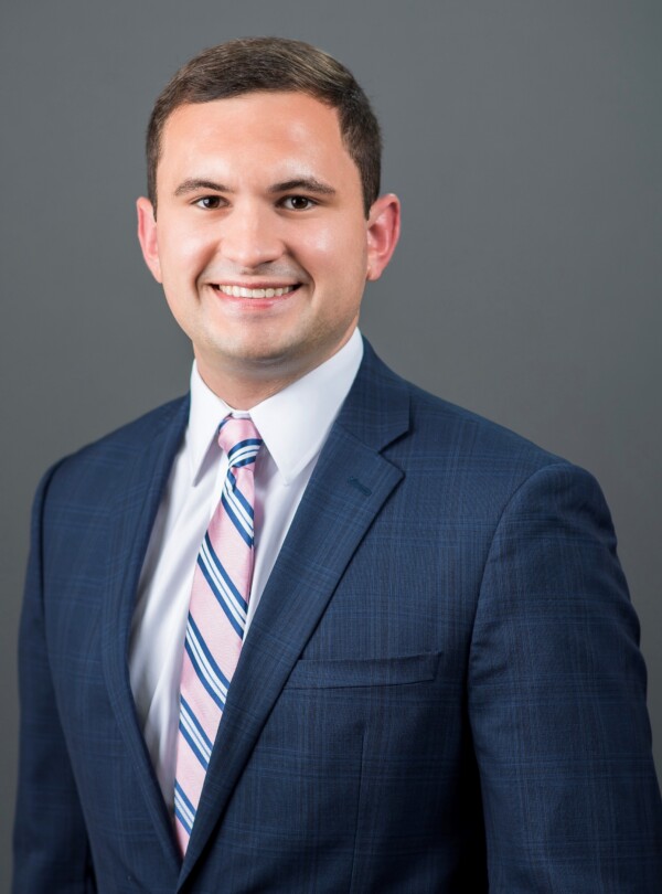 A young man in a dark suit and light blue shirt, smiling confidently against a gray background.