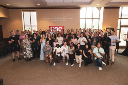 A large group of diverse individuals poses together in a hall, some making a hand gesture associated with the University of Southern California.