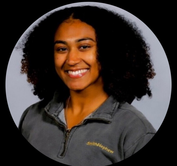 Smiling individual with curly hair wearing a gray quarter-zip top featuring a logo on the left side.