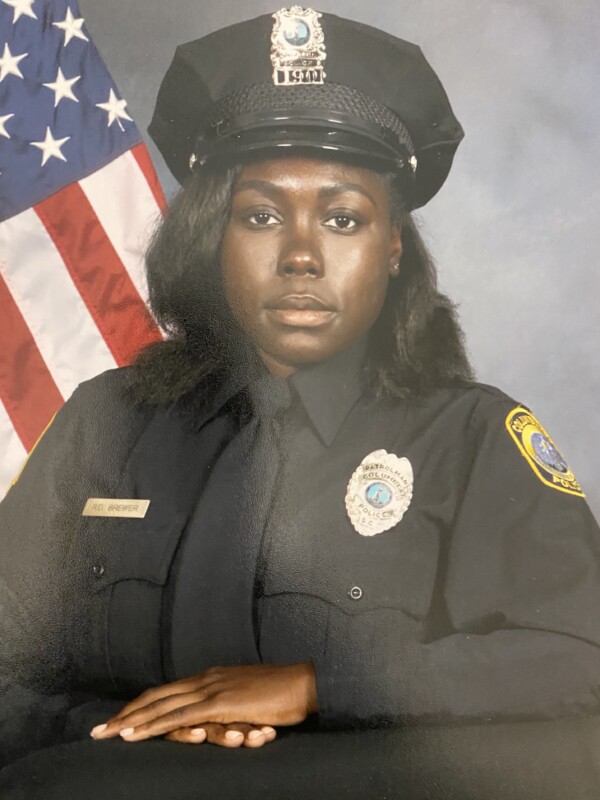 A police officer in uniform, seated confidently with a badge on her chest, against a backdrop featuring the American flag.