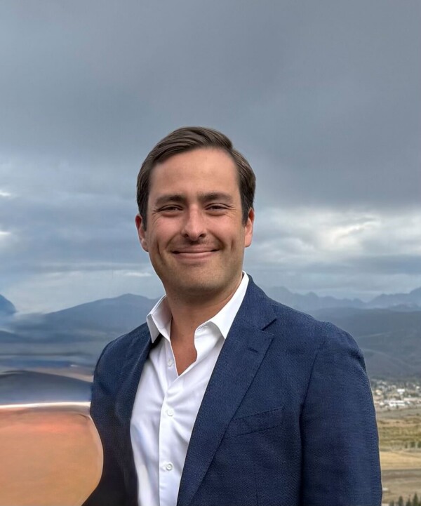 A man in a blue blazer and white shirt smiles against a backdrop of mountains and cloudy sky.