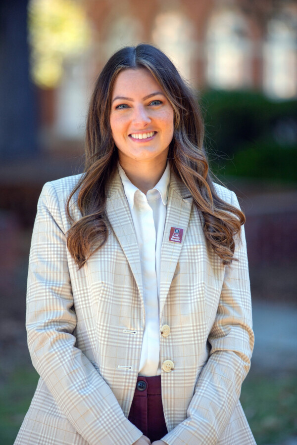 A woman with long wavy hair smiles confidently while wearing a light plaid blazer and white shirt, standing outdoors.
