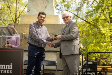 A young man in a checked shirt shakes hands with an older man in a suit, both smiling, in an outdoor setting near a monument.