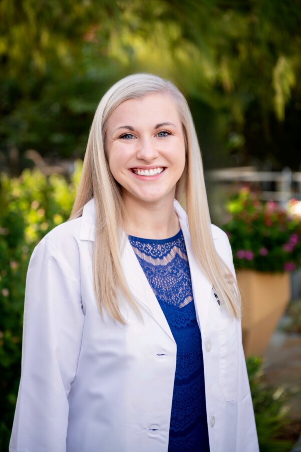 A smiling woman in a white lab coat, standing outdoors with greenery and flowers in the background.