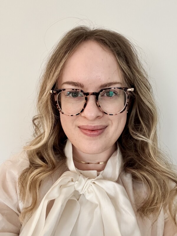 A smiling woman with long wavy blonde hair wearing tortoiseshell glasses and a cream-colored blouse with a bow tie.