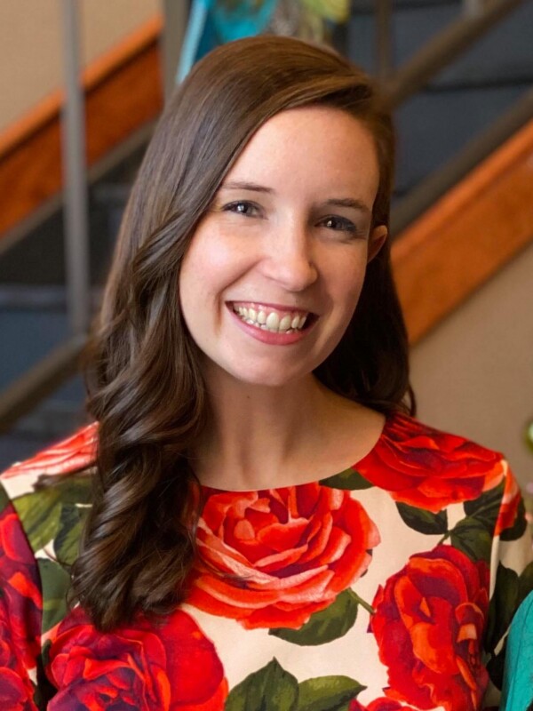 A woman with long brown hair smiles, wearing a colorful floral dress with large red roses, against a blurred background.