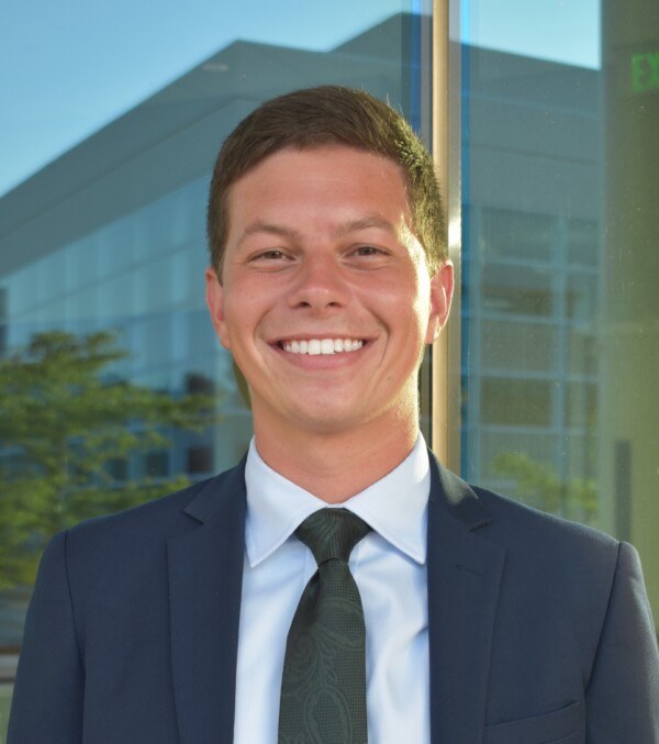 A smiling young man in a suit and tie stands outside, with a modern building and greenery visible in the background.