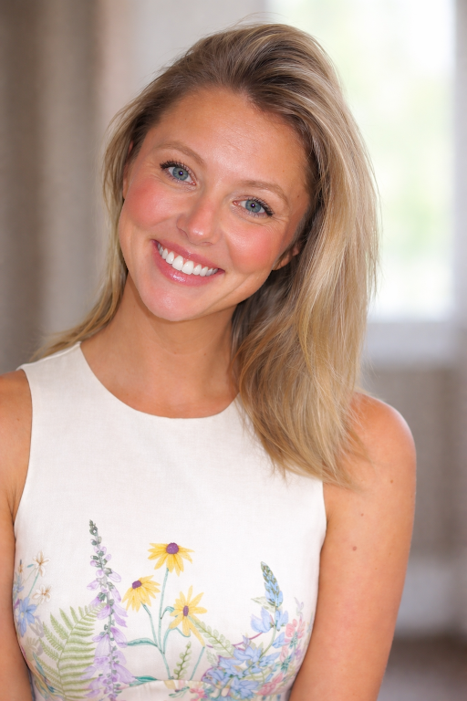 Smiling woman with long, wavy blonde hair, wearing a sleeveless floral dress, posing in a softly lit indoor setting.