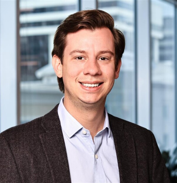 A smiling man in a blazer stands in front of large windows with a cityscape backdrop.