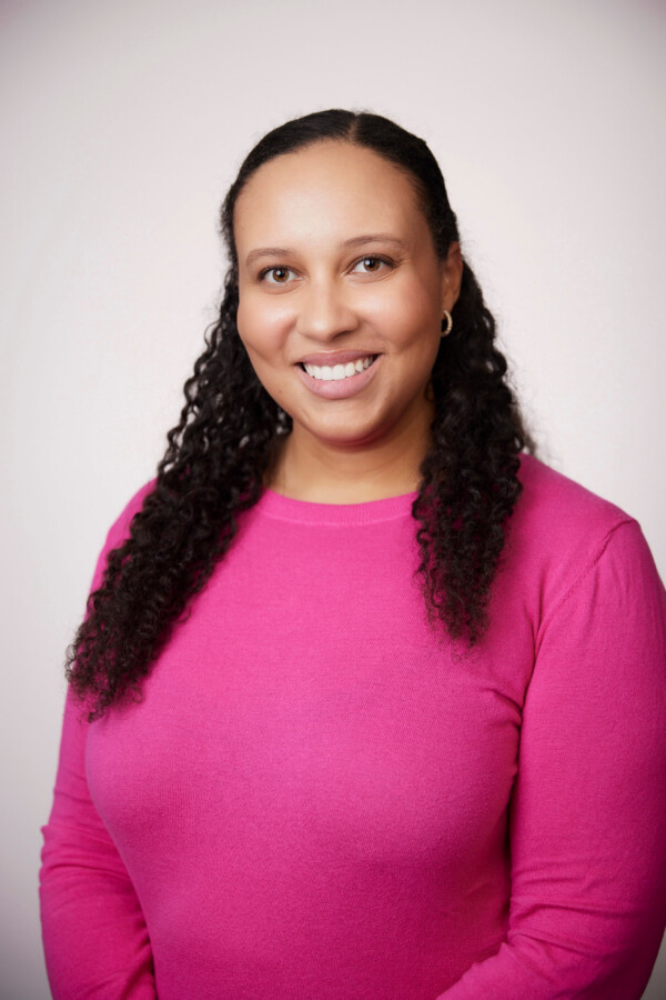 Smiling woman with curly hair wearing a pink sweater, standing against a light background.