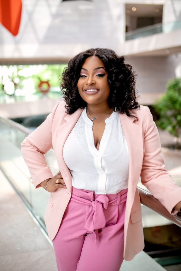 A smiling woman with curly hair wears a light pink blazer and white blouse, standing confidently in a modern, well-lit indoor space.