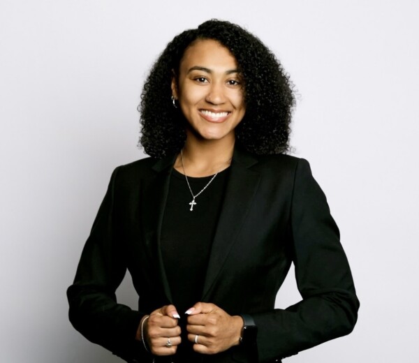 A woman with curly hair smiles while wearing a black blazer and a black shirt, with a cross necklace visible.