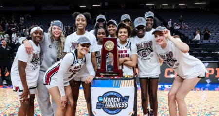 A group of female basketball players celebrating a championship win, posing with a trophy and wearing matching t-shirts in a festive atmosphere.