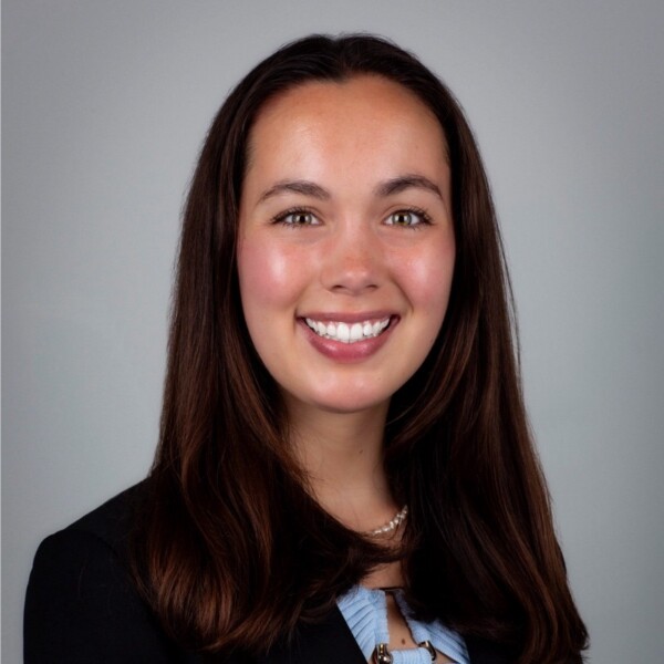 A smiling woman with long, dark hair and a professional outfit, against a gray background. She has a pearl necklace and a friendly expression.