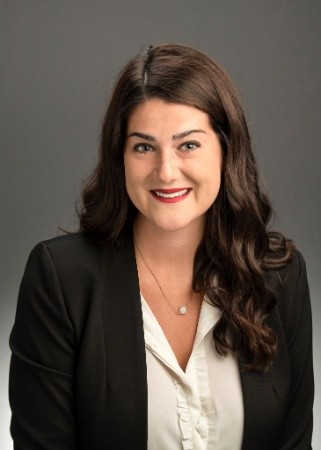 A woman with long, dark, wavy hair smiles at the camera, wearing a black blazer over a white blouse.