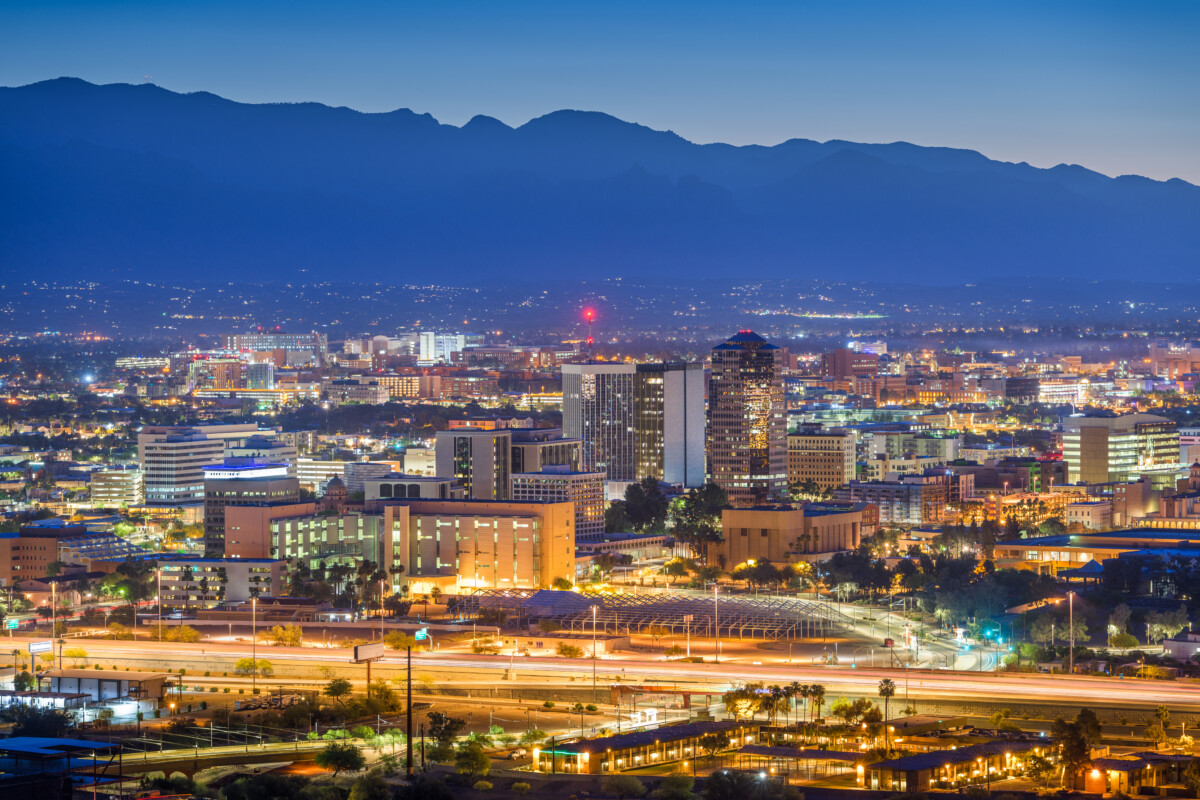 City skyline at dusk with glowing lights from buildings and highways, set against distant mountains.
