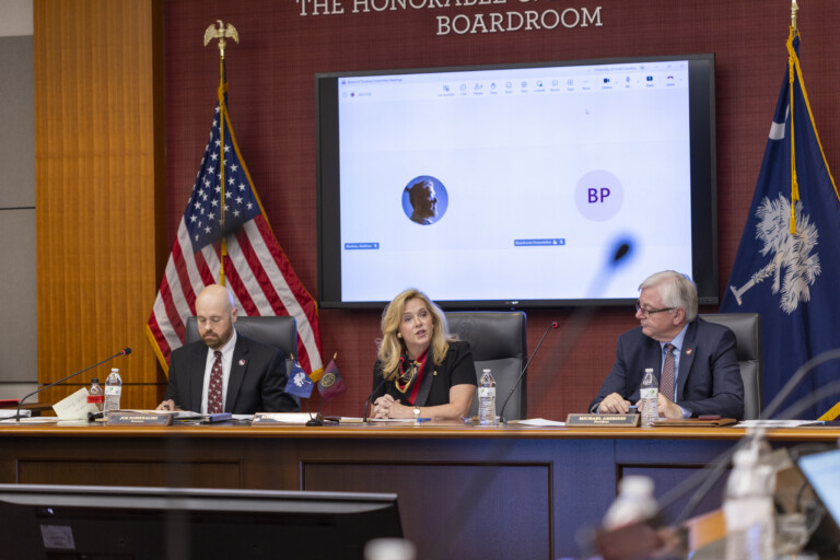 Three officials seated at boardroom table during formal presentation with American flag backdrop.