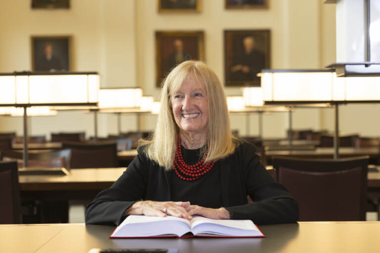 A smiling woman with blonde hair wearing a black blazer and red beads sits at a desk with an open book in a library.