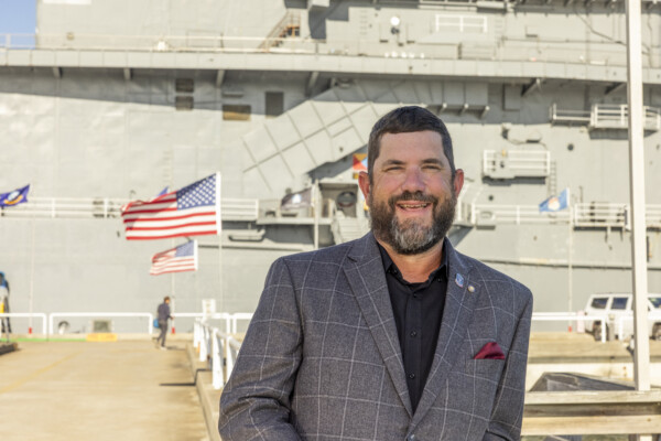 Man in blazer smiling on naval ship dock with American flags visible behind him